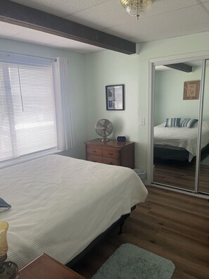 Ein Schlafzimmer mit einem Bett, einem Schminktisch, einem Nachttisch, einem Ventilator, einer Uhr, einem Fenster mit Jalousien und einem Spiegel.