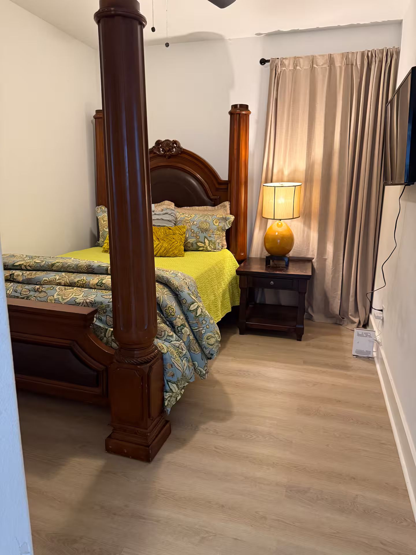 Een slaapkamer met een hemelbed, een nachtkastje met een lamp en een raam met gordijnen.