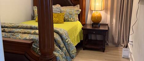 A bedroom with a four-poster bed, a bedside table with a lamp, and a window with curtains.