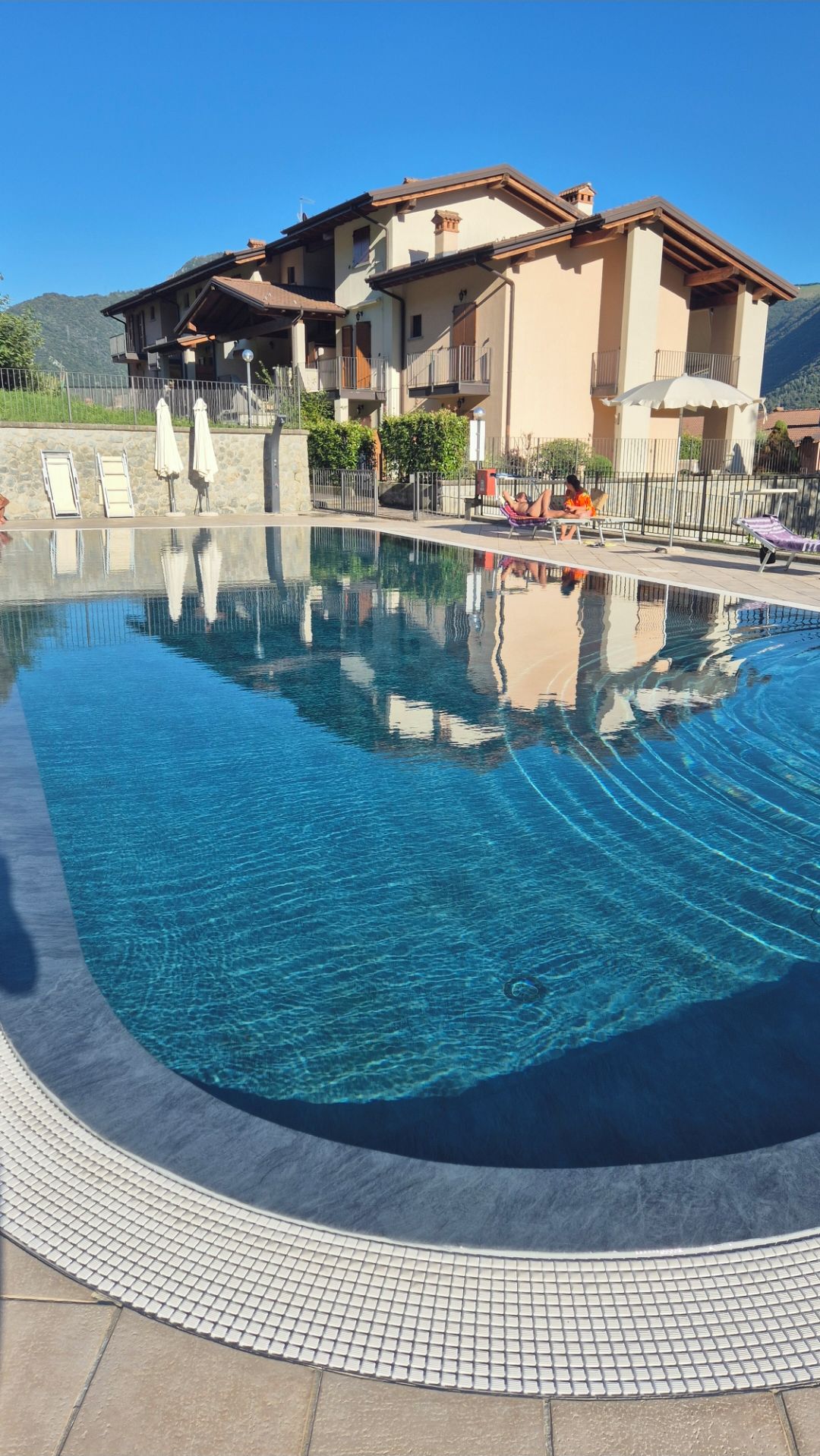 A swimming pool with a tiled edge, surrounded by lounge chairs and umbrellas, with a building in the background.