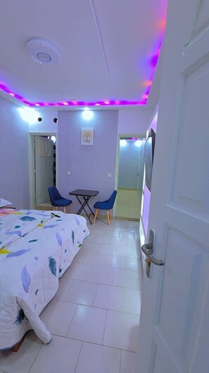 Ein Schlafzimmer mit einem Bett, zwei Stühlen, einem kleinen Tisch, einer Tür und einer Deckenleuchte.