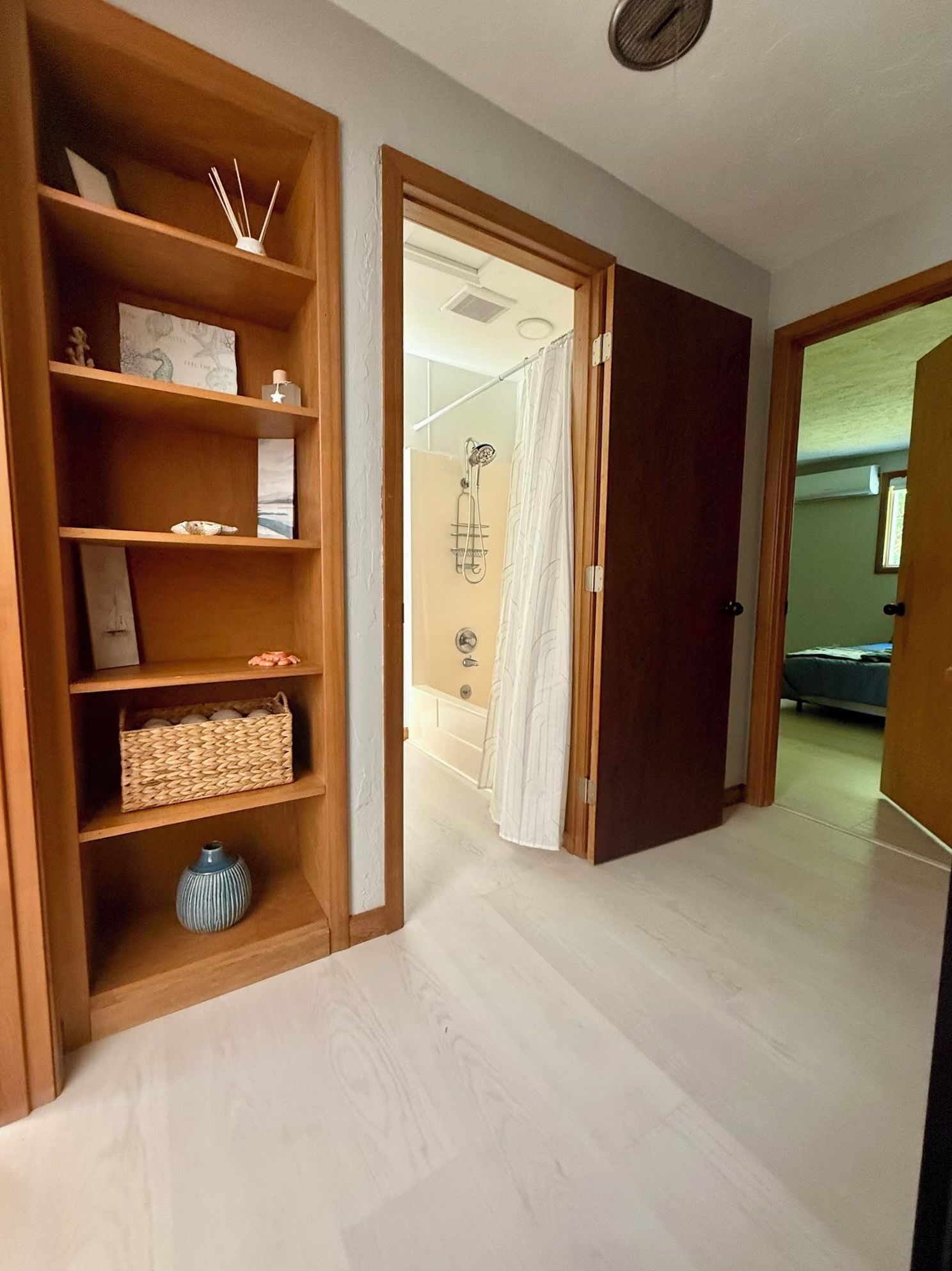A hallway with a wooden bookshelf, a shower area, and a door leading to another room.