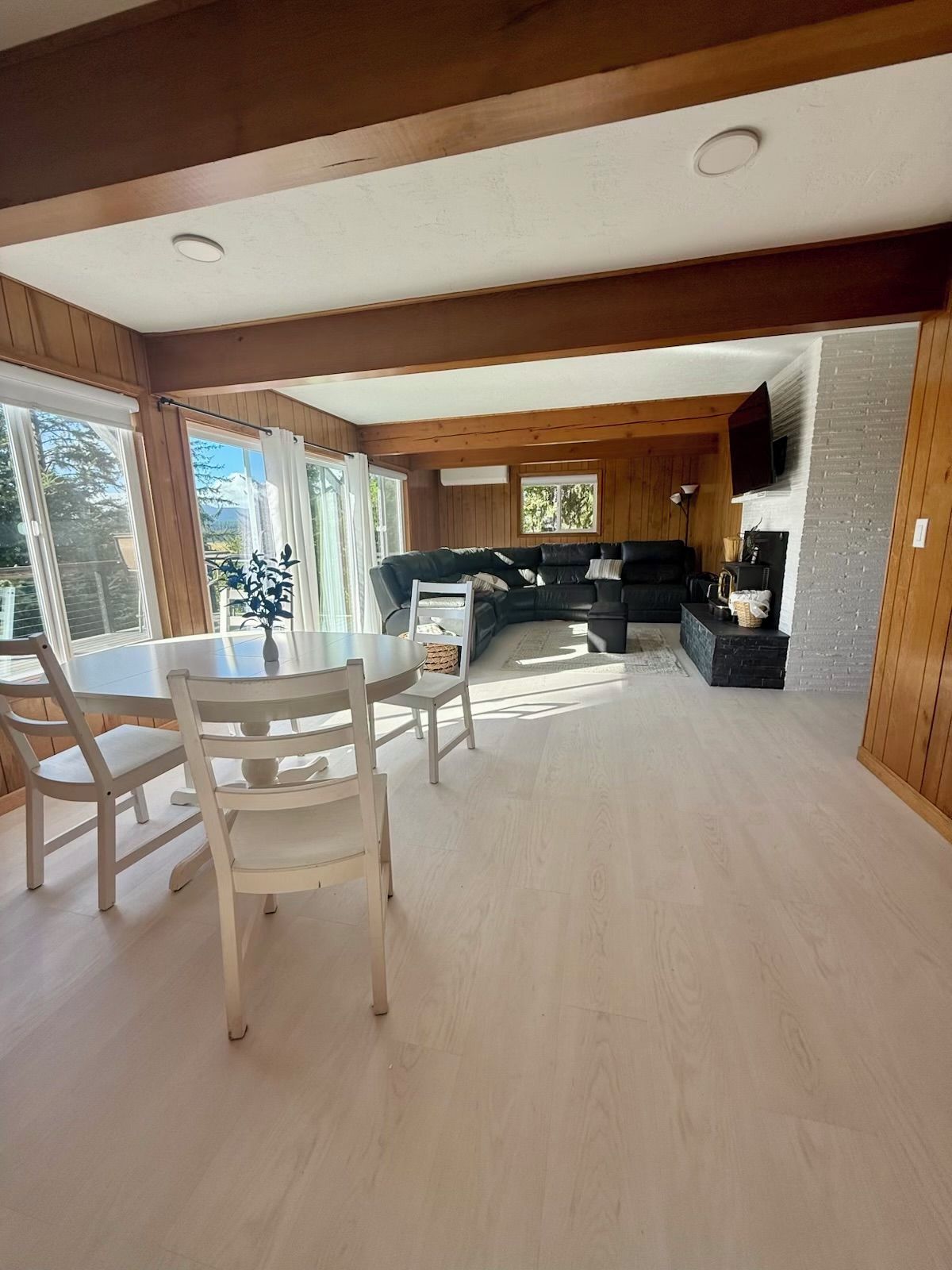 A spacious living area with a dining table and chairs, a black sofa, and a television mounted on a brick wall.