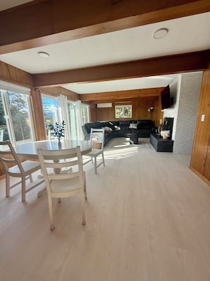 A spacious living area with a dining table and chairs, a black sofa, and a television mounted on a brick wall.