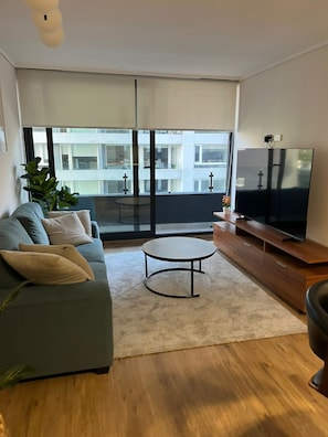 A modern living room with a sofa, a coffee table, a TV on a wooden stand, and a balcony with a view of buildings.