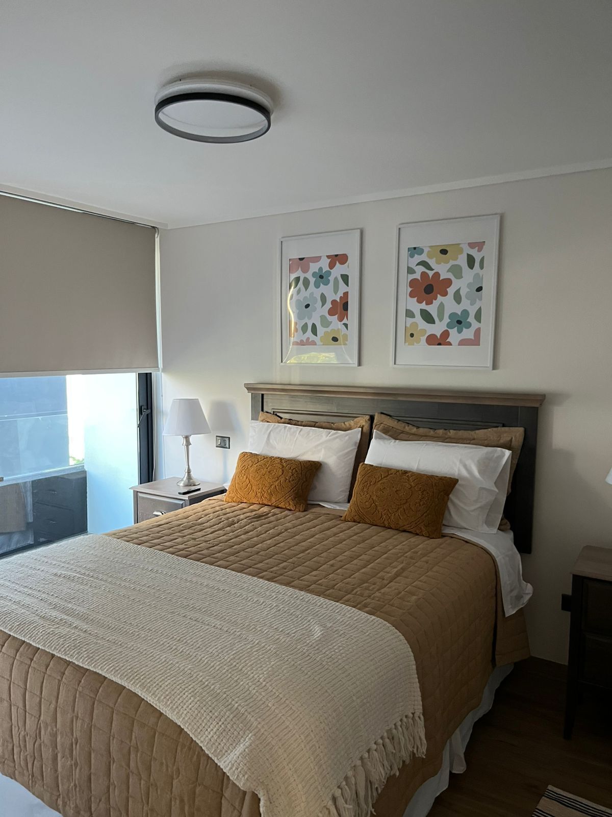 Une chambre d’hôtel avec un lit, une table de chevet avec une lampe, une fenêtre donnant sur un paysage agréable et deux œuvres d’art florales encadrées accrochées au mur.