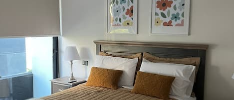 A hotel room with a bed, a bedside table with a lamp, a window with a view, and two framed floral artworks on the wall.