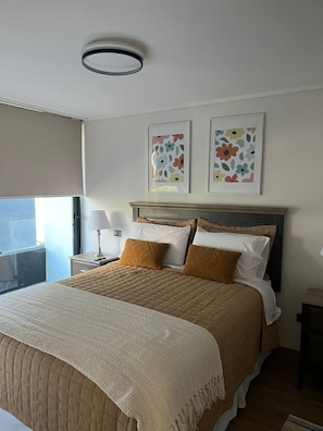 Une chambre d’hôtel avec un lit, une table de chevet avec une lampe, une fenêtre donnant sur un paysage agréable et deux œuvres d’art florales encadrées accrochées au mur.
