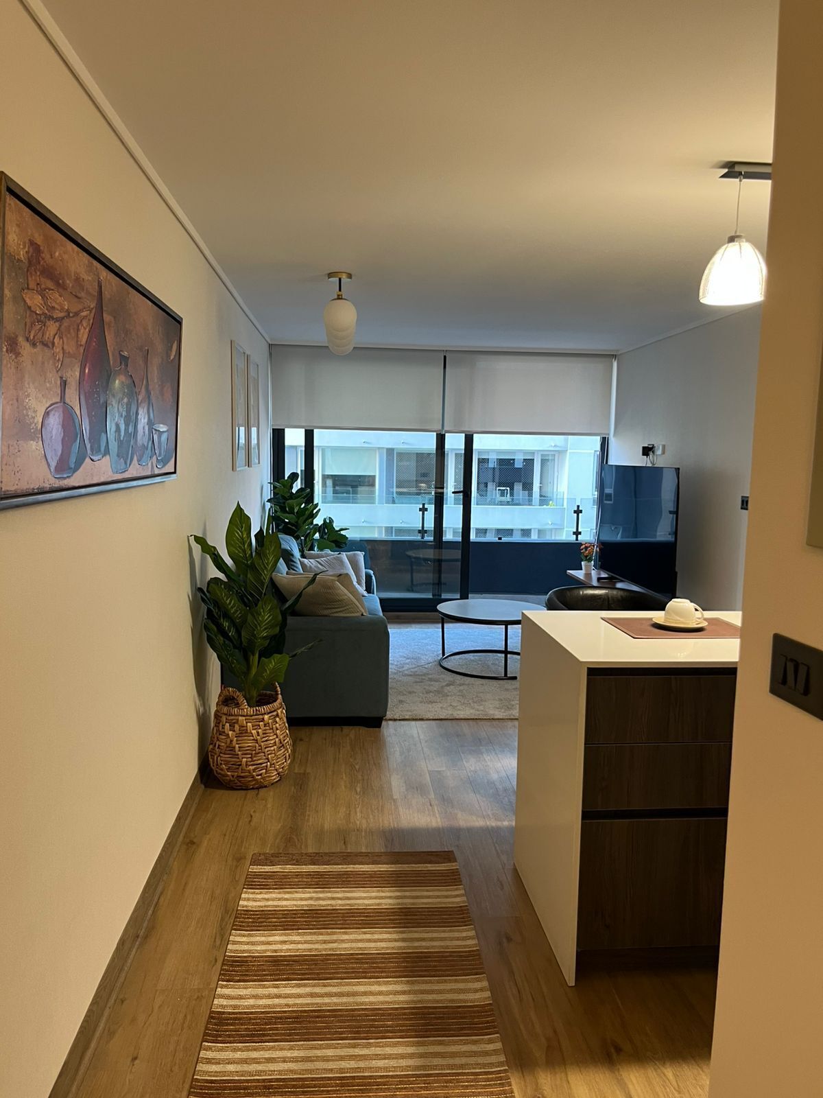 Un appartement moderne comprenant un salon, une cuisine et un coin repas. Il y a une grande fenêtre, un tableau au mur et une plante en pot.