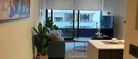 A modern apartment with a living room, kitchen, and dining area. There is a large window, a painting on the wall, and a potted plant.