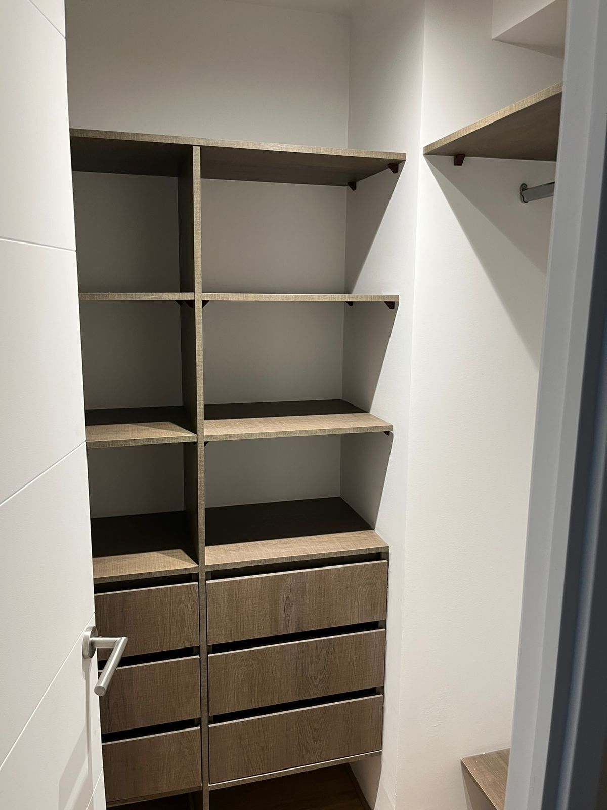 Un armario vestidor con varias estanterías y cajones, una puerta con manija, y un interruptor de luz en la pared.