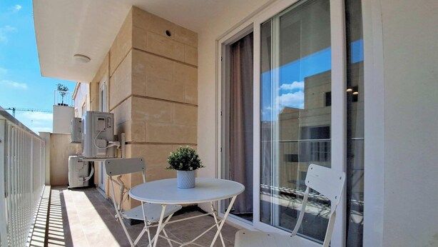 A balcony with a table and chairs, a potted plant, and a large window. - Gozo Stay: 2BR Apt with Balcony in Xagħra Village (Ix-Xagħra)