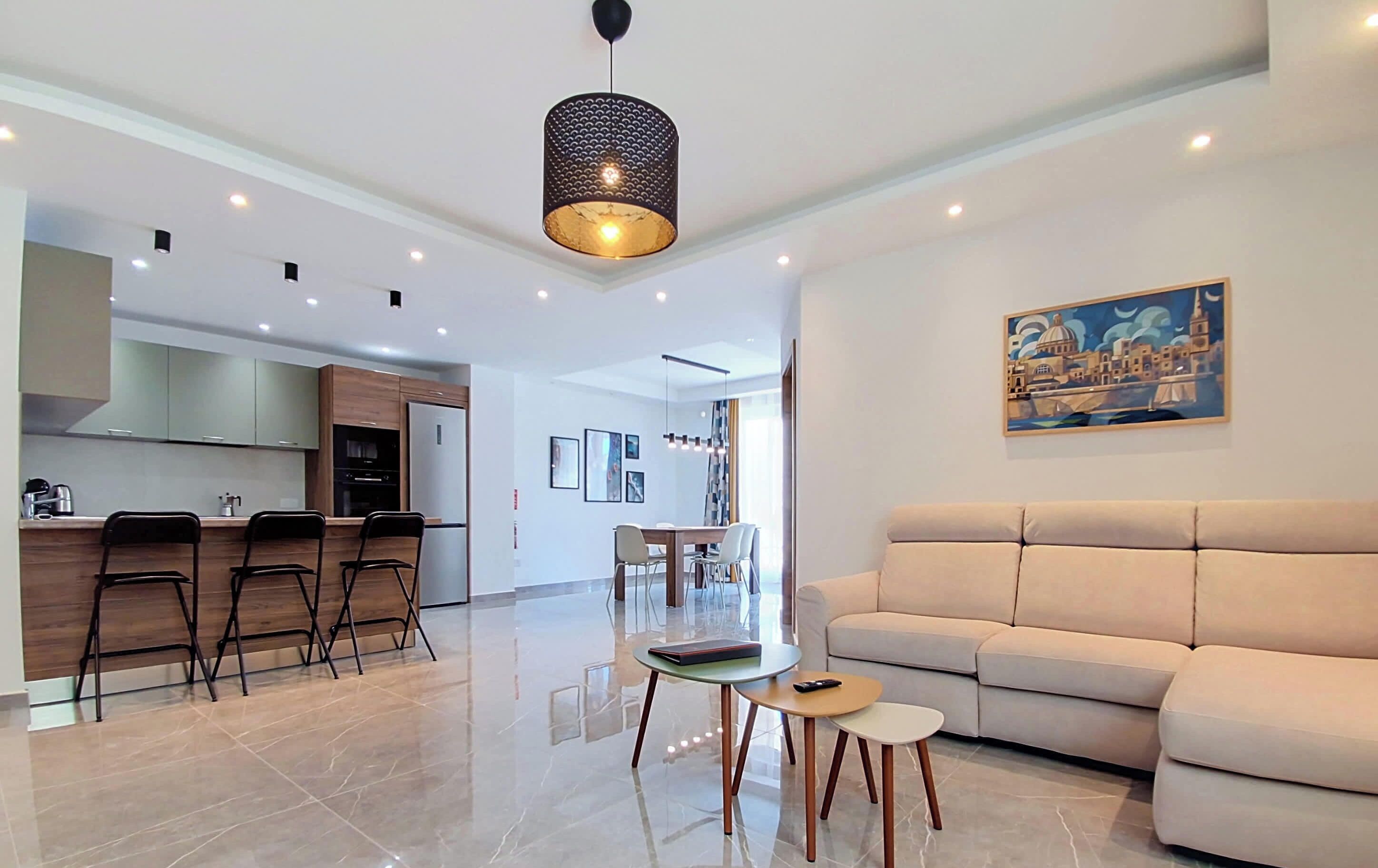Una sala moderna con un sofá, un comedor con sillas, una cocina con taburetes de bar y una lámpara colgante.