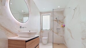 A modern bathroom with a marble wall, a round mirror, a sink, a toilet, and a glass-enclosed shower area. - Gozo Stay: 2BR Apt with Balcony in Xagħra Village (Ix-Xagħra)