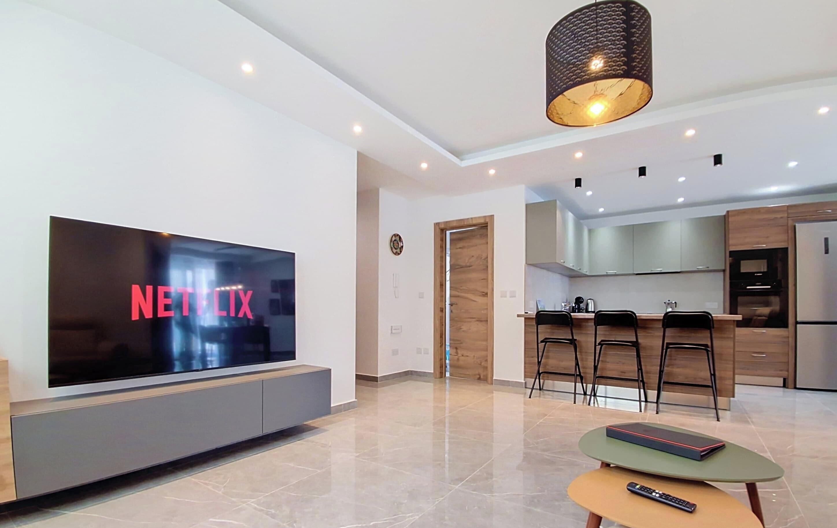 Una sala de estar moderna con un televisor de pantalla plana, una mesa de centro y una zona de cocina con taburetes de bar.