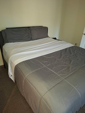 Ein Bett mit einer grau-weißen Tagesdecke, einem dunklen Kopfteil und einem Teppichboden.