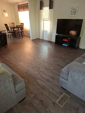 Una sala de estar con piso de madera, un sofá gris, un área de comedor con sillas, un mueble para TV con televisor de pantalla plana y una pared con una cita motivacional.