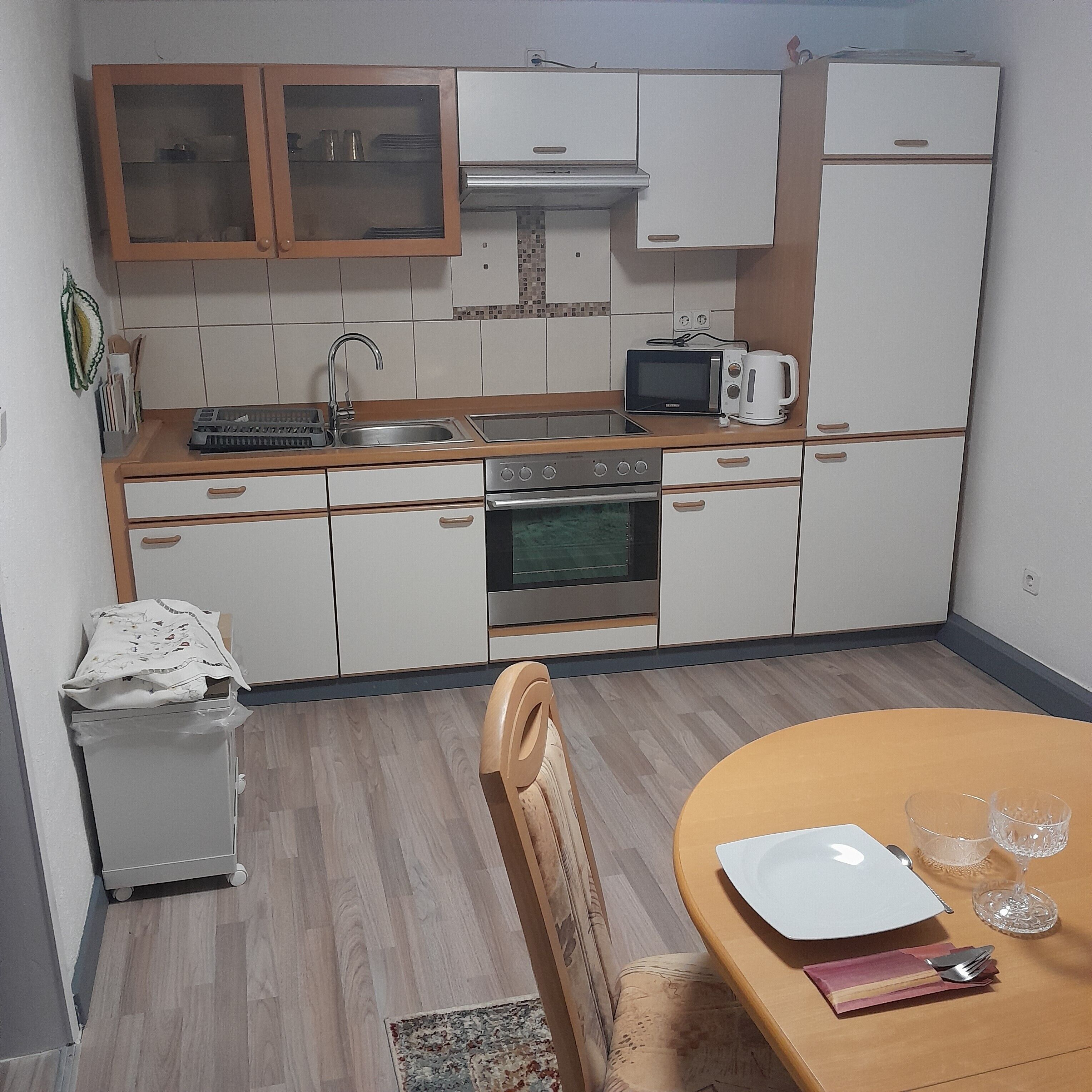 Een compacte keuken met een magnetron, broodrooster en oven. Er is een klein spoelgedeelte en een kast met glazen deuren. Een eettafel met een stoel is zichtbaar.