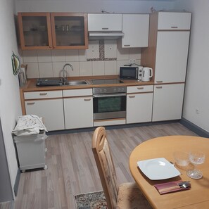 A compact kitchen with a microwave, toaster, and oven. There is a small sink and a cabinet with glass doors. A dining table with a chair is visible.