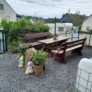 Eine Terrasse mit Holztisch und Bänken, umgeben von Kies, Blumentöpfen und einer Steinstatue.