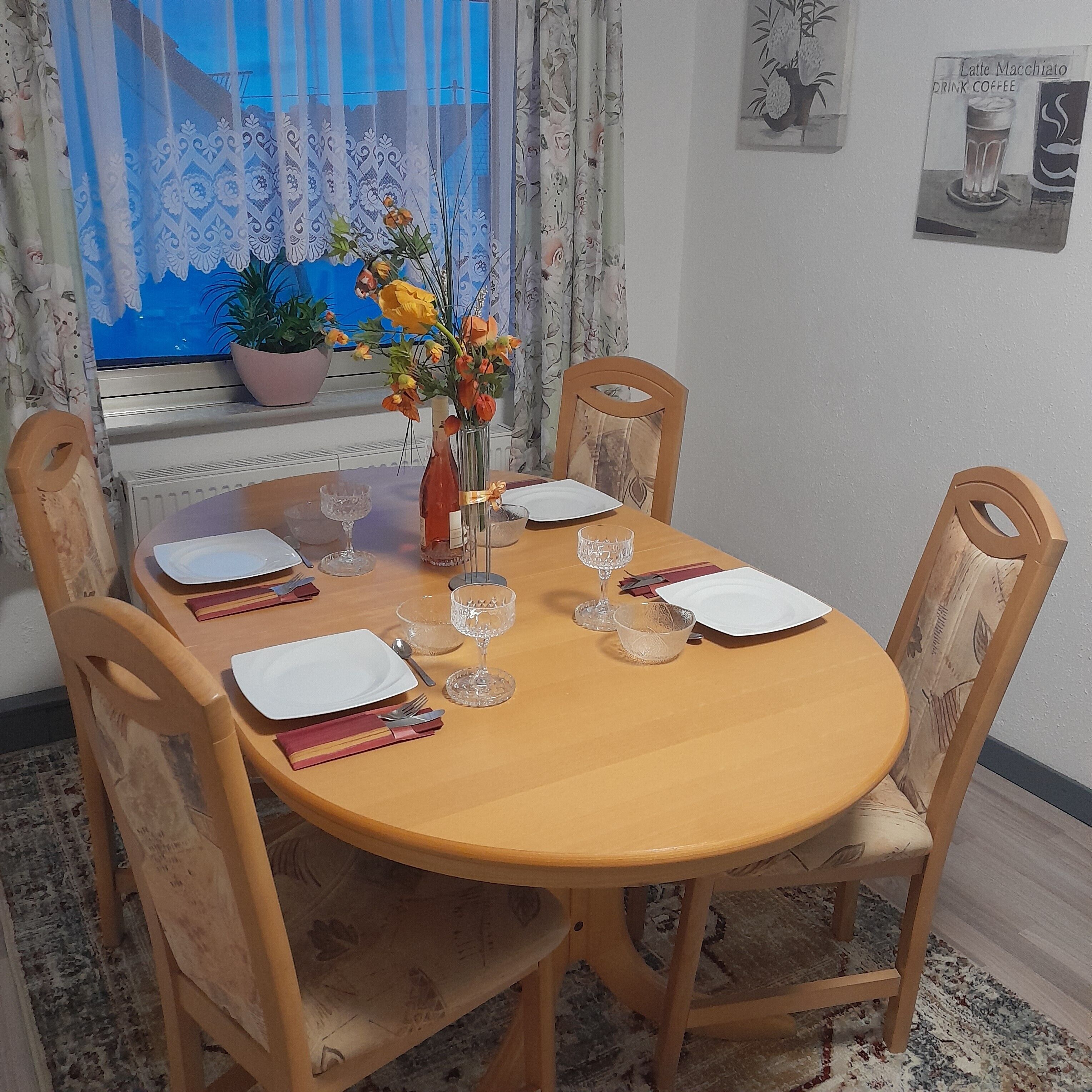 A dining table set for four with chairs, a vase with flowers, and a window with curtains.