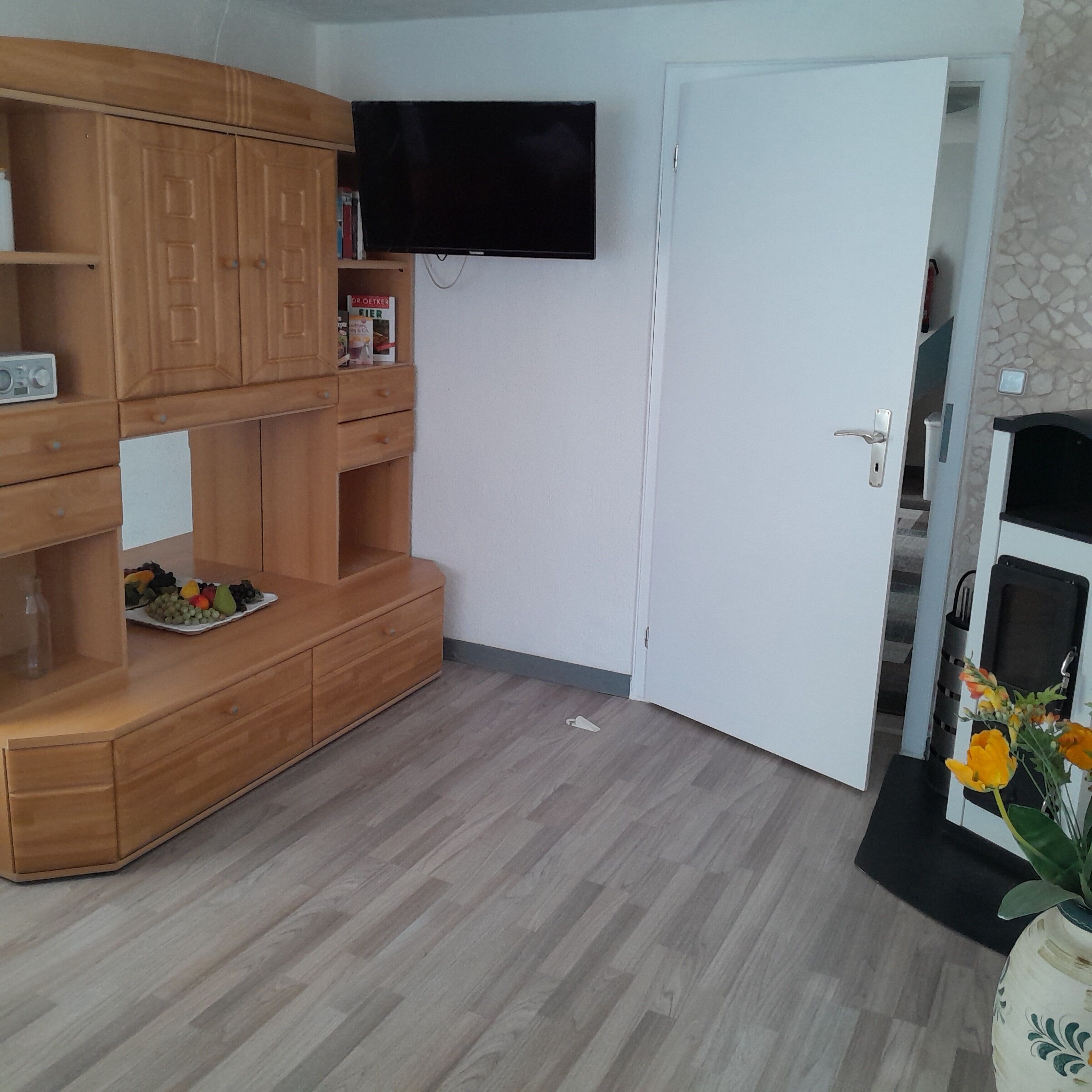A room with a wooden cabinet, a flat-screen TV mounted on the wall, a white door, and a small plant with yellow flowers.
