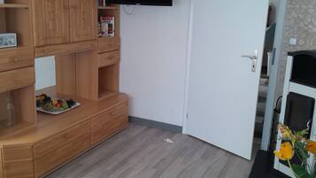 A room with a wooden cabinet, a flat-screen TV mounted on the wall, a white door, and a small plant with yellow flowers.