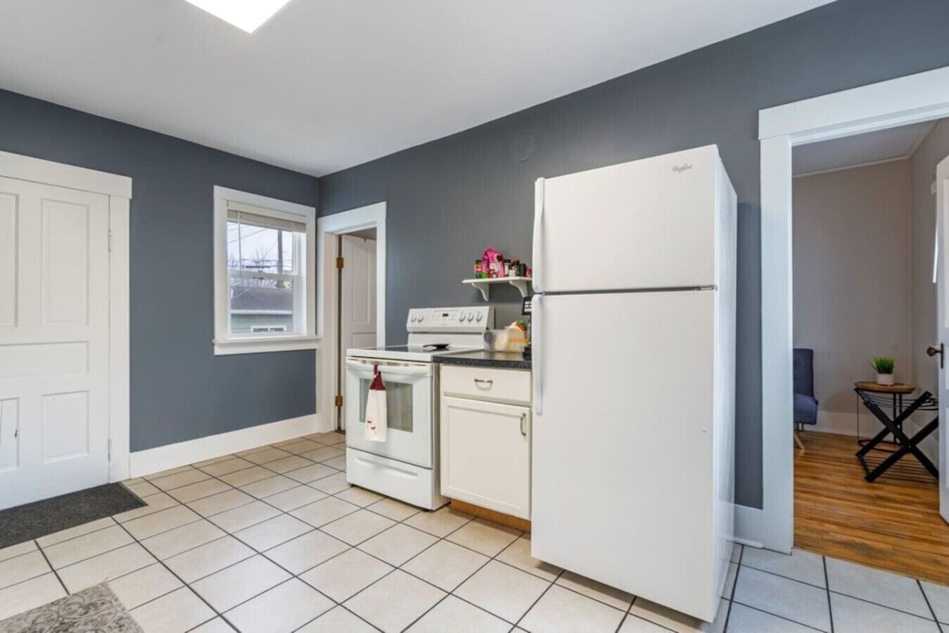 A compact kitchen with white appliances, a window, and a door leading to another room.