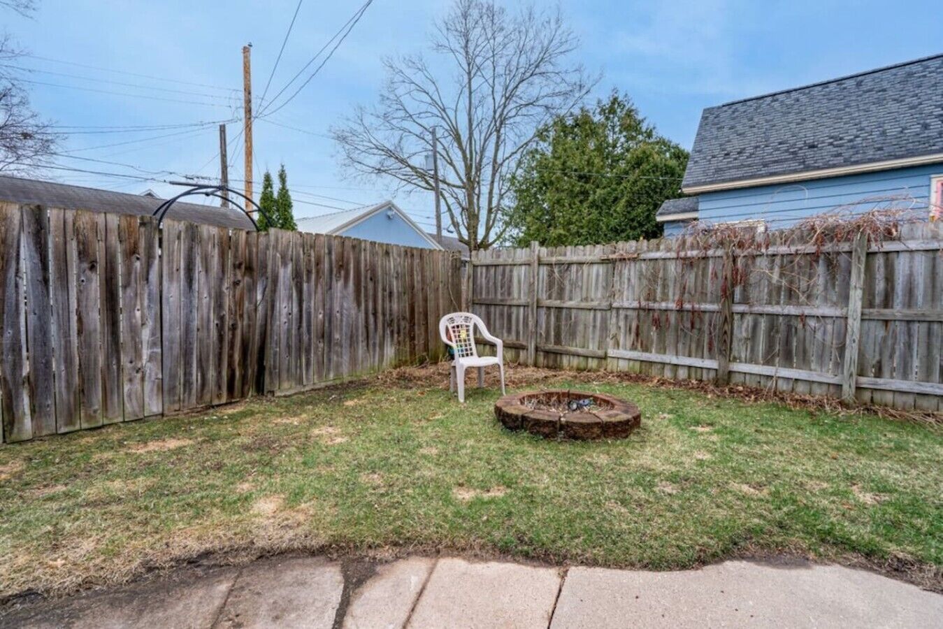 A backyard with a wooden fence, a white plastic chair, and a fire pit.