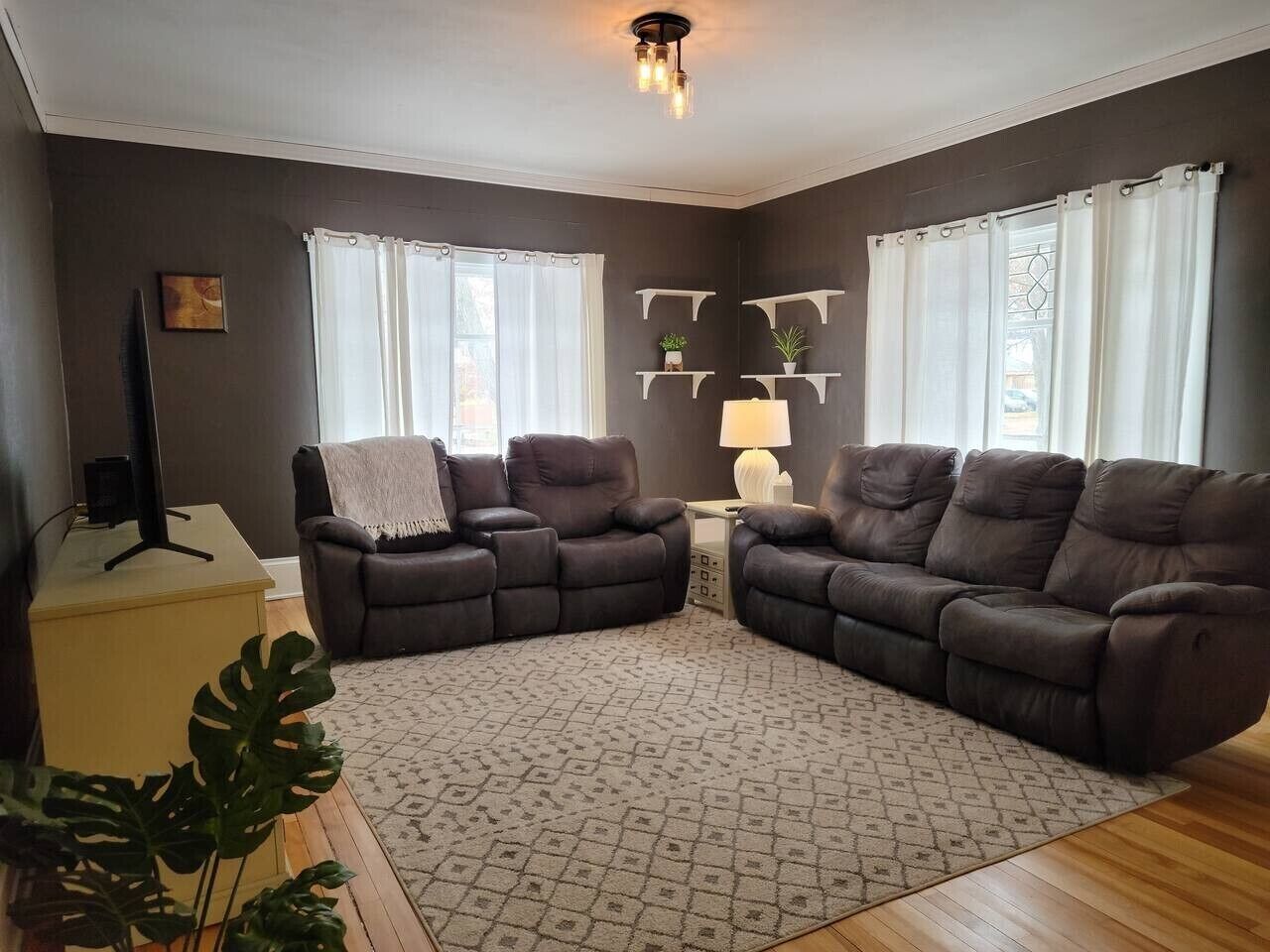 Ein Wohnzimmer mit zwei Sofas, einem Couchtisch und einer Lampe. Zwei Fenster mit Vorhängen, eine Pflanze und ein an der Wand befestigtes Regal sind ebenfalls zu sehen.