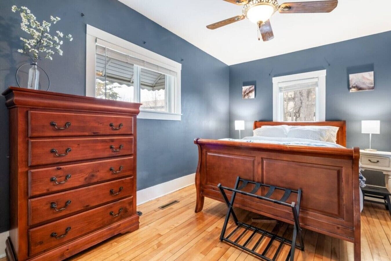 Une chambre avec une commode en bois, un lit, un ventilateur de plafond et deux fenêtres.