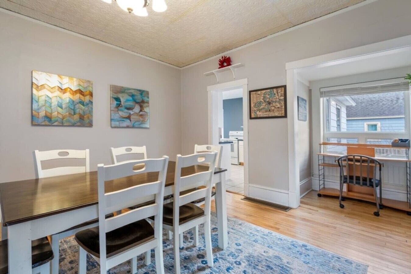 Un coin repas avec une table en bois et des chaises blanches, un tapis à motifs bleus et des œuvres d’art accrochées aux murs.