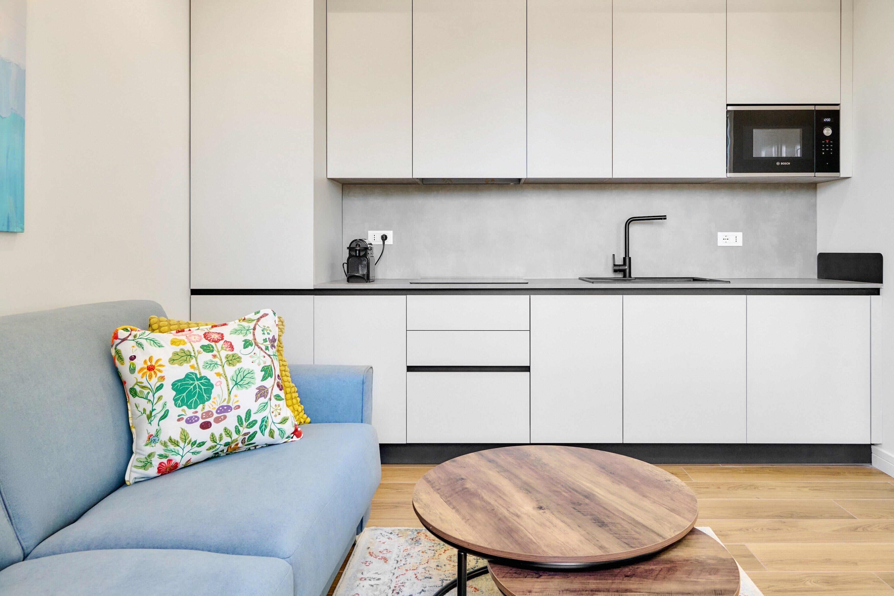 A modern kitchen with white cabinets, a Bosch microwave, a sink, and a blue sofa with a colorful pillow.