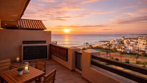 Ein Balkon mit Blick auf den Sonnenuntergang über dem Meer, ein Tisch mit Stühlen und ein kleiner Topfpflanze.