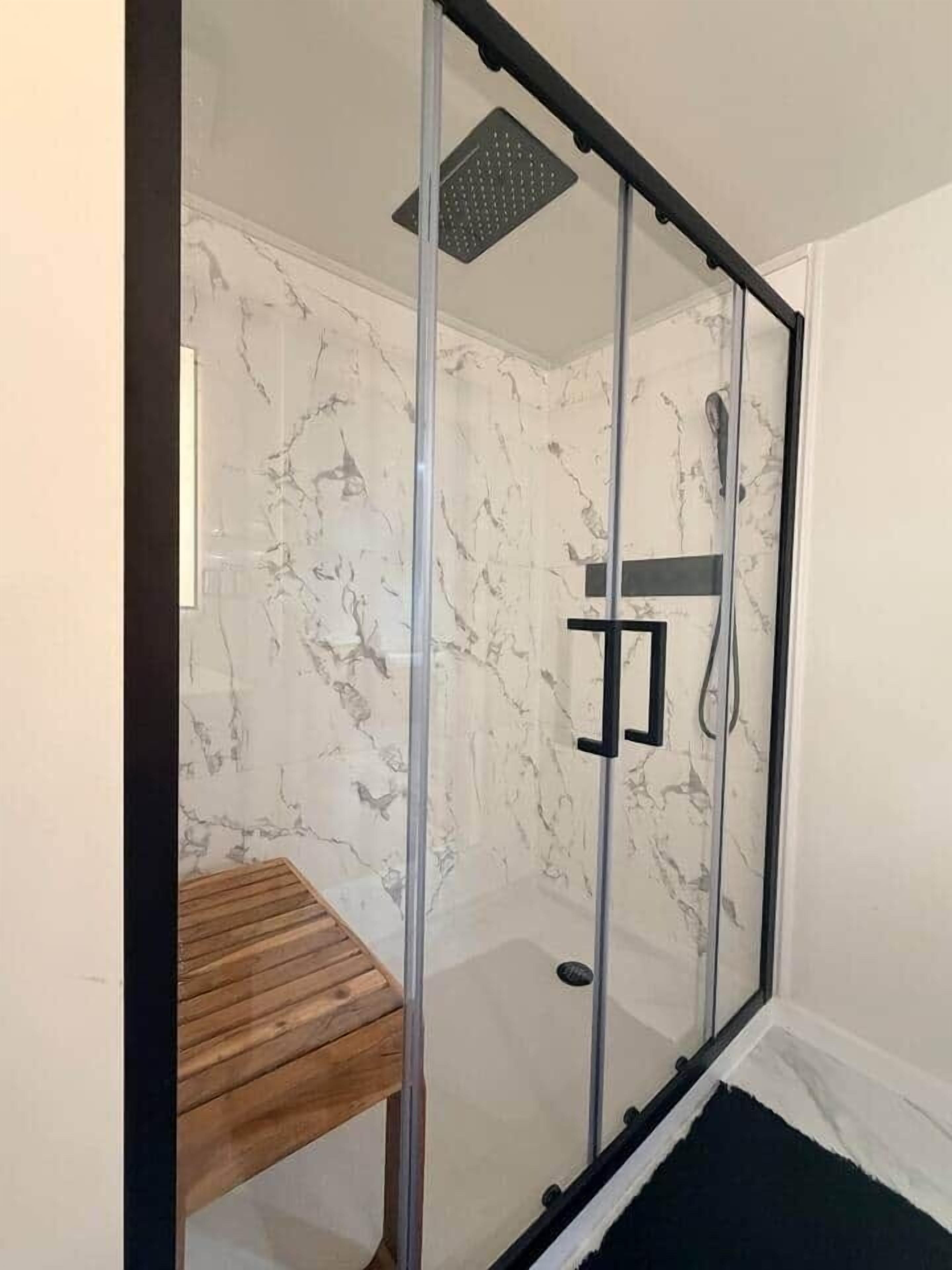 A glass-enclosed shower with a marble wall, a wooden bench, and a black mat.