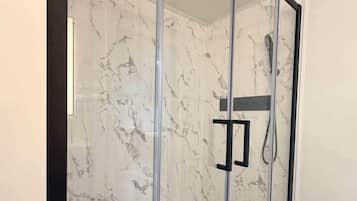 A glass-enclosed shower with a marble wall, a wooden bench, and a black mat.
