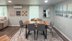 A dining area with a wooden table, grey chairs, and a rug. There are framed pictures on the walls and an air conditioning unit above the table.