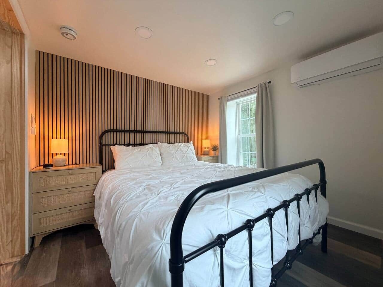 A bedroom with a bed, bedside tables, a window, and a wall-mounted air conditioner.