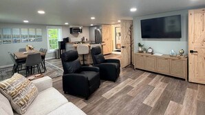 A modern living room with a sofa, two black armchairs, a dining area with a table and chairs, a kitchen with appliances, and a TV mounted on the wall.