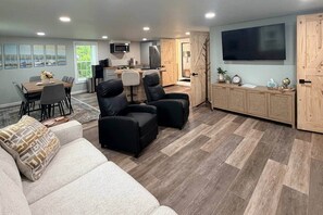 Ein modernes Wohnzimmer mit einer Couch, zwei schwarzen Sesseln, einer Essecke mit Tisch und Stühlen, einer Küche mit Geräten und einem an der Wand befestigten Fernseher.