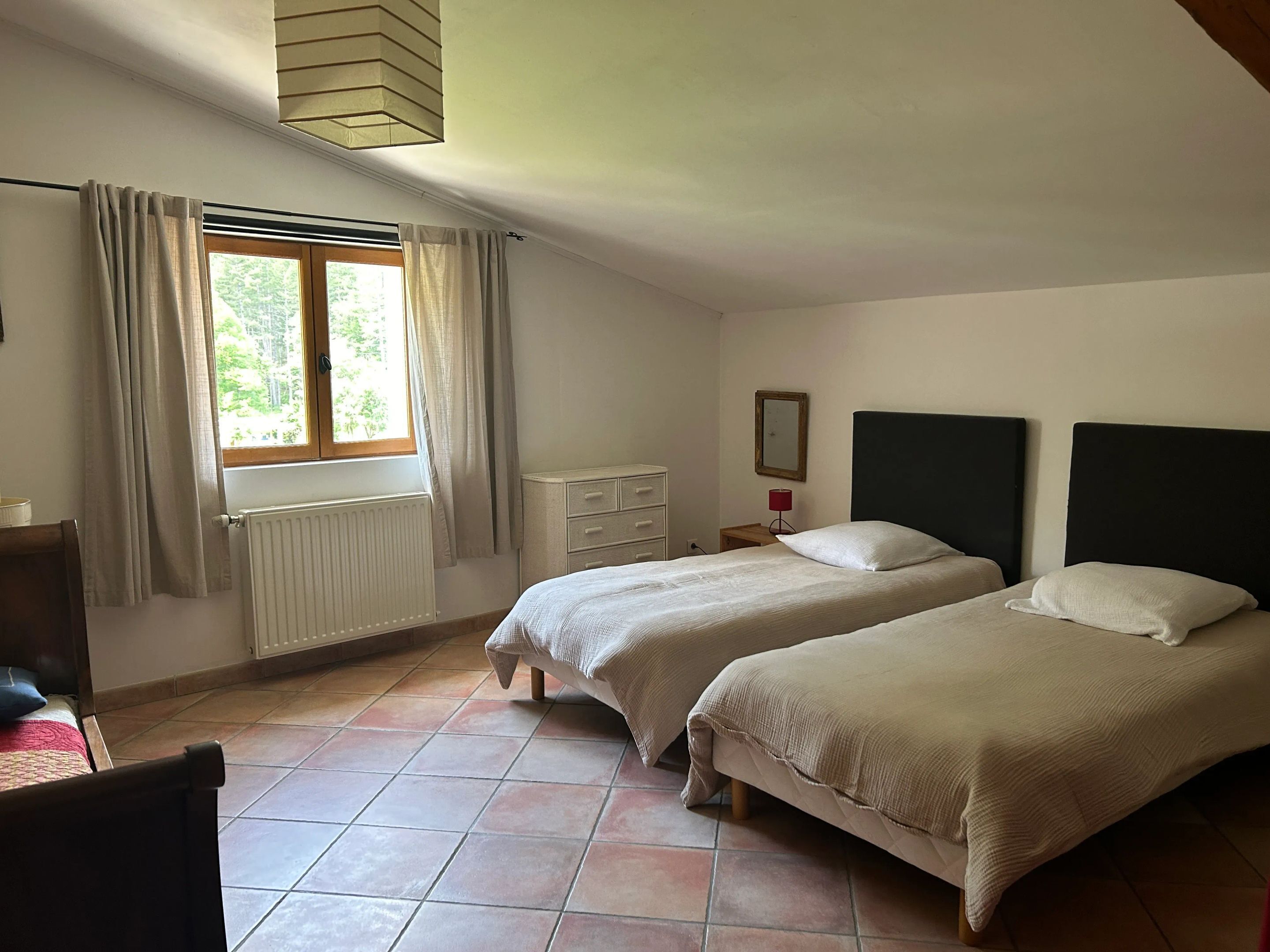 Een slaapkamer met twee bedden, een radiator, een raam met gordijnen, een nachtkastje met een lamp en een spiegel aan de muur.