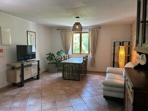 Ein Wohnzimmer mit Fernseher, Esstisch, Stühlen, einer Couch, einer Zimmerpflanze und einem Fenster mit Vorhängen.