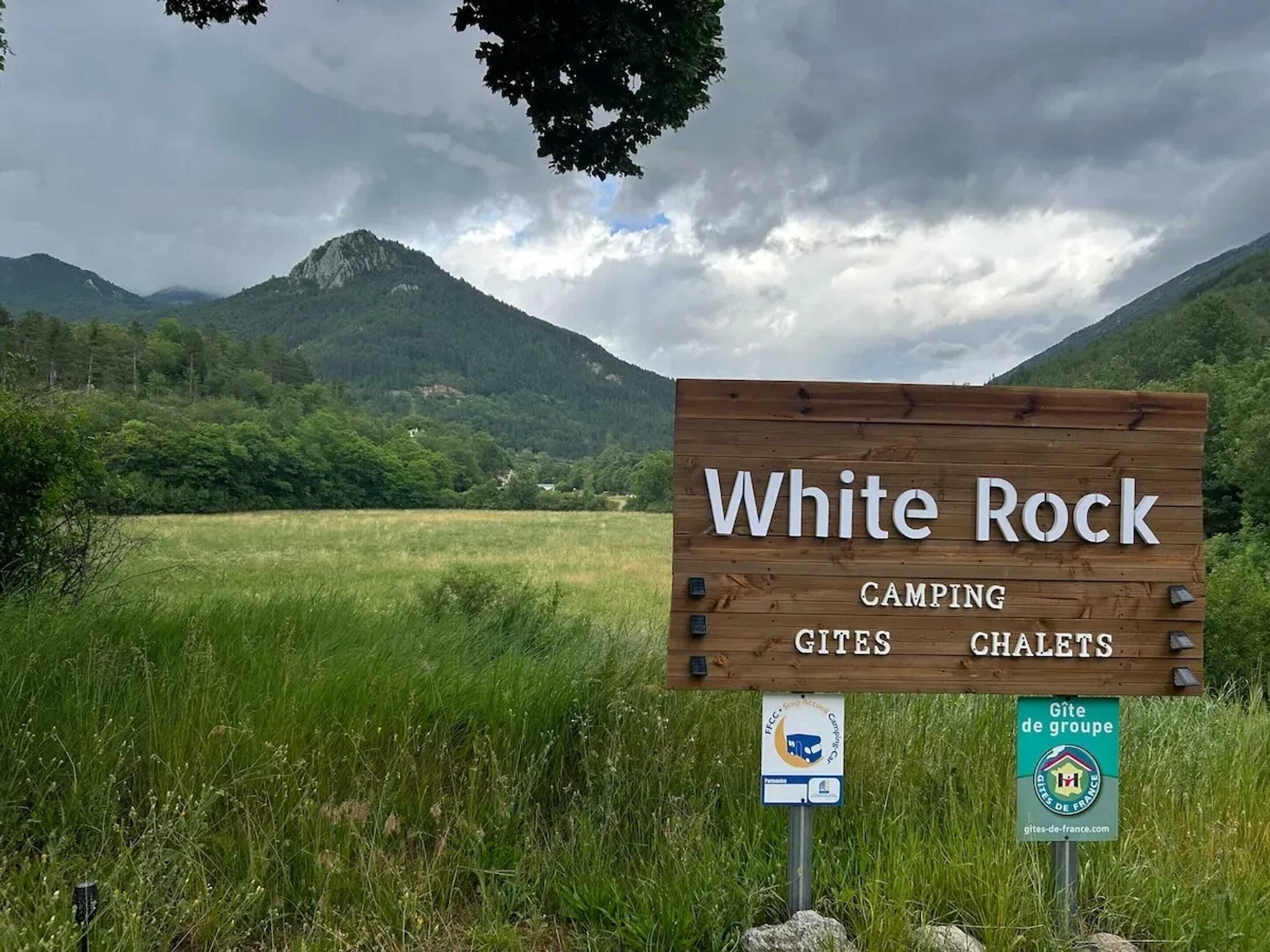 Een houten bord met daarop 'White Rock' en 'Camping Gites Chalets', met op de achtergrond een grasveld en bergen.