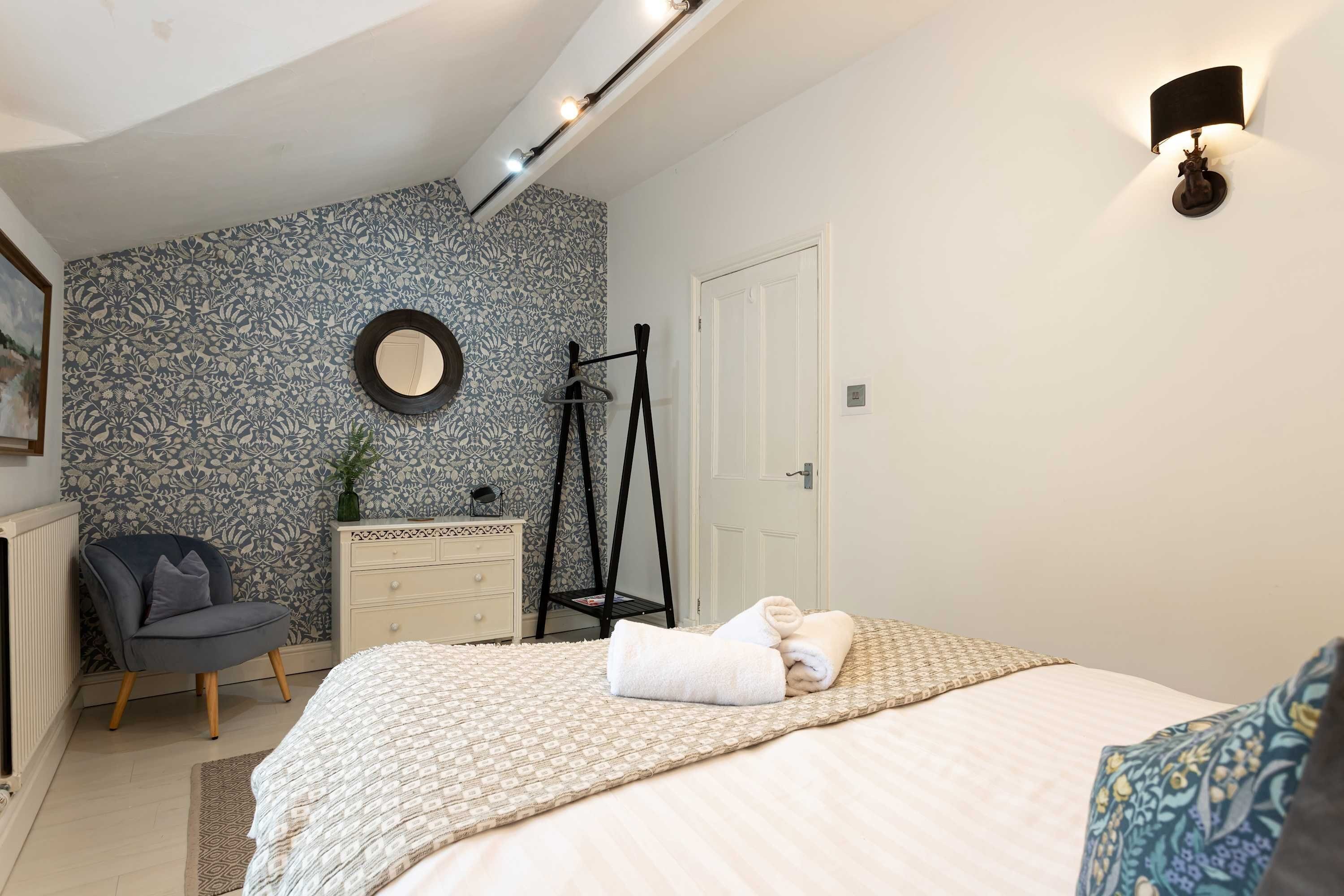 Un dormitorio con cama, una silla, cómoda, espejo y lámpara de pared.