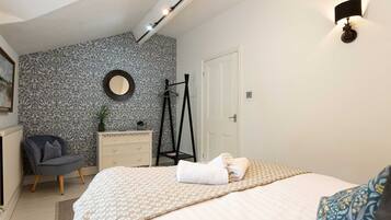 Un dormitorio con cama, una silla, cómoda, espejo y lámpara de pared.