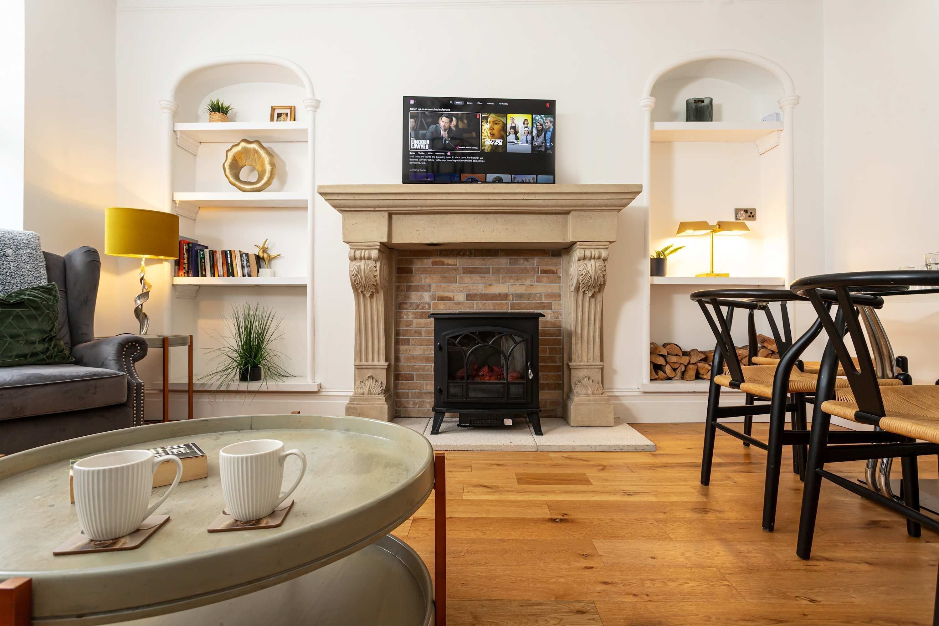 A living room with a fireplace, a television mounted above it, a sofa, a coffee table with cups, and a dining area with chairs.