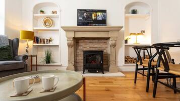 A living room with a fireplace, a television mounted above it, a sofa, a coffee table with cups, and a dining area with chairs.