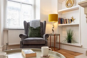 Eine gemütliche Leseecke mit einem Ledersessel, einem kleinen Tisch mit einem Buch und einer Tasse, einer Lampe, einem Bücherregal mit Büchern und einem dekorativen Gegenstand sowie einer Topfpflanze.