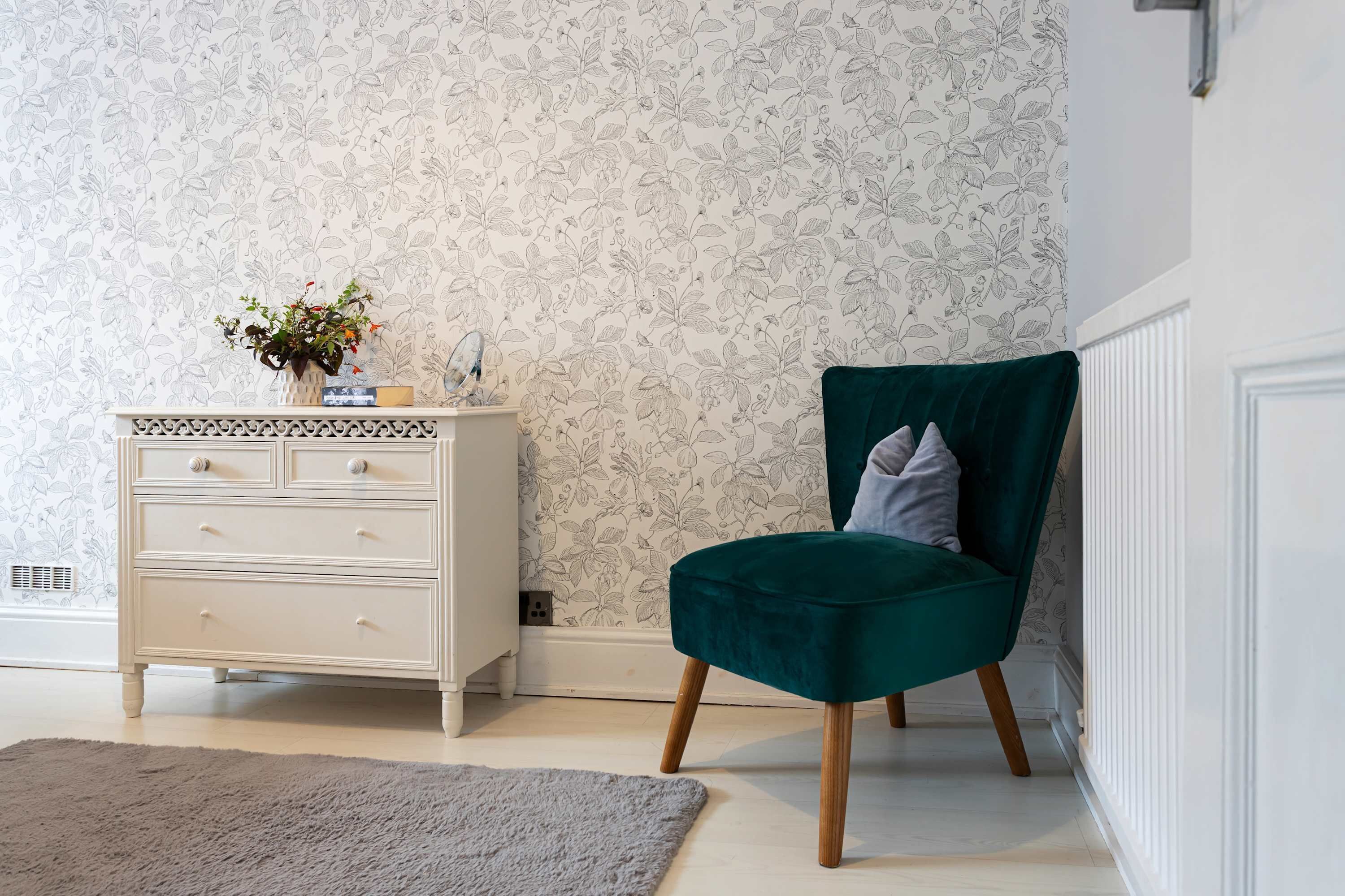 Una habitación con un aparador blanco, un sillón de terciopelo verde y un papel tapiz floral.