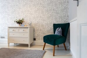 Ein Zimmer mit einem weißen Schminktisch, einem grünen Samtstuhl und floralem Wandpapier.